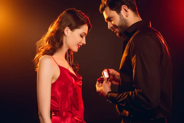Side view of man holding gift box with jewelry ring near smiling woman in red dress on black background with lighting — Stock Photo