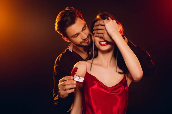 Handsome man covering girlfriend eyes and holding gift box with jewelry ring on black background with lighting — Stock Photo