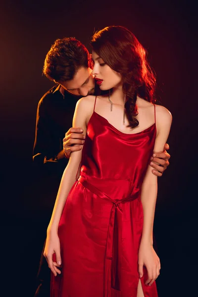 Handsome man kissing shoulder of elegant girlfriend in red dress isolated on black — Stock Photo