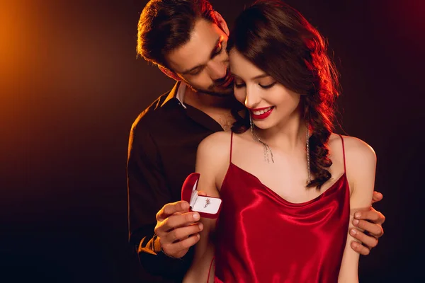 Hombre guapo sosteniendo caja de regalo con anillo de joyería y abrazo novia sonriente sobre fondo negro con iluminación - foto de stock