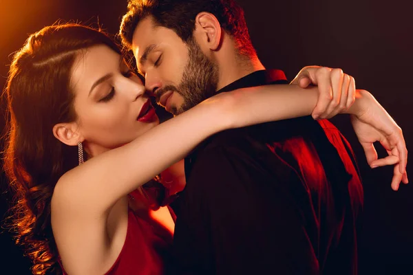 Vista lateral do homem bonito beijando namorada elegante com lábios vermelhos no fundo preto com iluminação — Fotografia de Stock