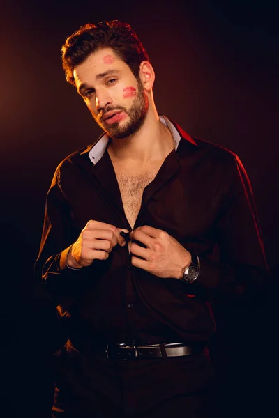 Handsome man with lipstick prints on face looking at camera and unbuttoning shirt isolated on black — Stock Photo