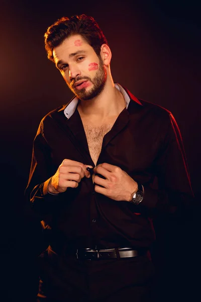 Hombre guapo con impresiones de lápiz labial en la cara mirando a la cámara y desabotonando la camisa aislada en negro - foto de stock