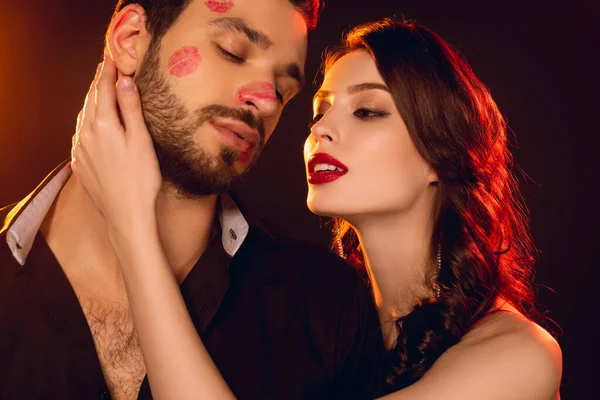 Attractive girl touching handsome boyfriend with lipstick prints on face on black background — Stock Photo