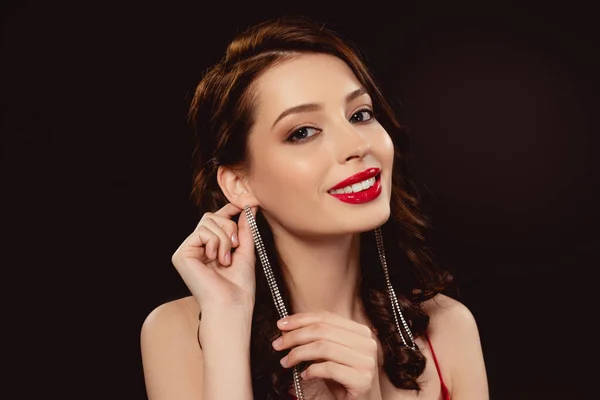 Beautiful woman with red lips touching earring and smiling at camera isolated on black — Stock Photo