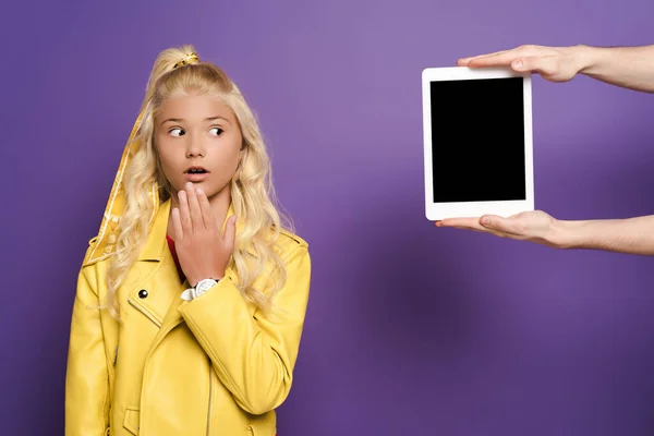 Cropped view of man giving digital tablet to shocked kid on purple background — Stock Photo