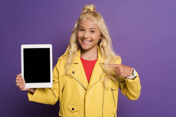 Garoto sorrindo apontando com o dedo para tablet digital em fundo roxo — Fotografia de Stock