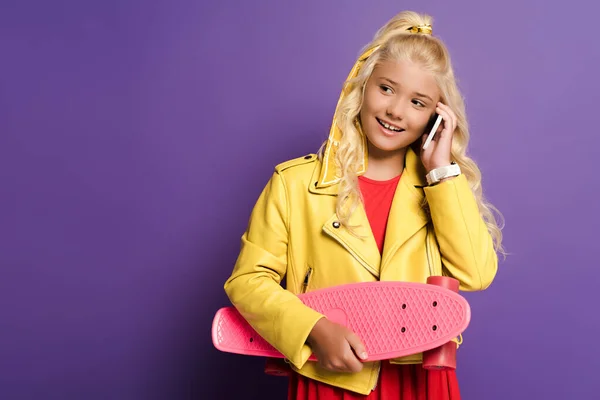 Smiling kid holding penny board and talking on smartphone on purple background — Stock Photo