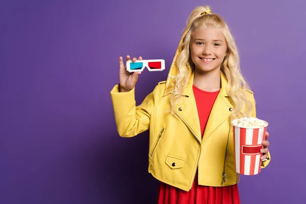 Smiling kid holding 3d glasses and bucket with popcorn on purple background — Stock Photo