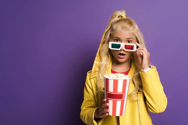 Schockiertes Kind mit 3D-Brille, das Eimer mit Popcorn auf lila Hintergrund hält — Stockfoto