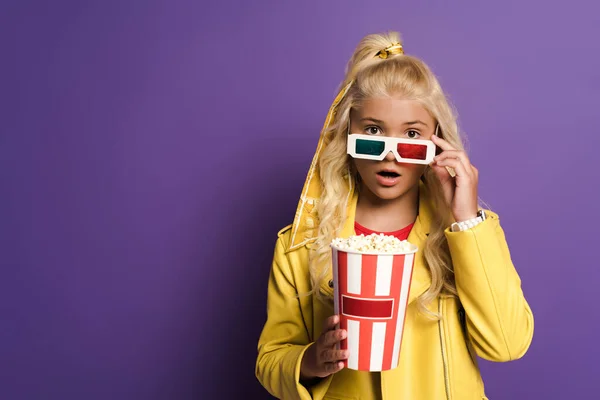 Enfant choqué avec des lunettes 3D tenant seau avec pop-corn sur fond violet — Photo de stock