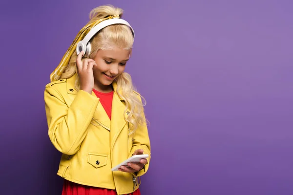 Enfant souriant avec écouteurs utilisant un smartphone sur fond violet — Photo de stock