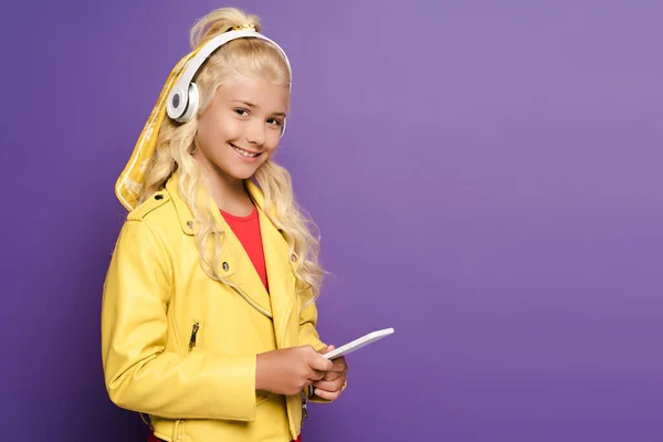 Smiling kid with headphones holding smartphone on purple background — Stock Photo