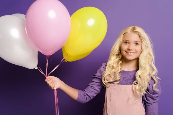 Niño sonriente sosteniendo globos y mirando a la cámara sobre fondo púrpura - foto de stock