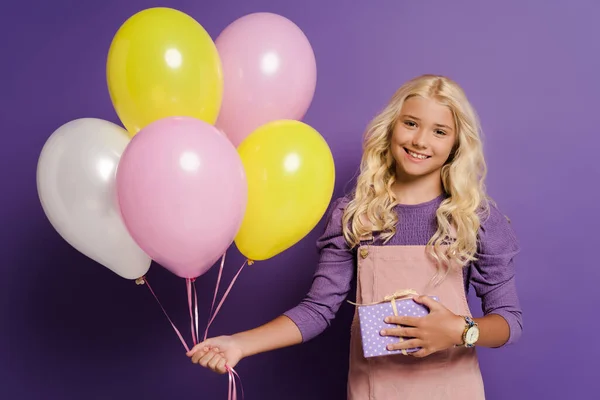 Criança sorrindo segurando balões e caixa de presente no fundo roxo — Fotografia de Stock