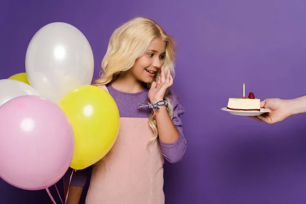Cropped view of woman giving plate with birthday cake to smiling kid with balloons on purple background — Stock Photo