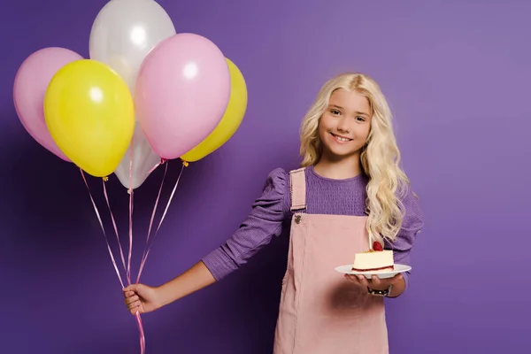 Niño sonriente sosteniendo globos y plato con pastel de cumpleaños sobre fondo morado - foto de stock