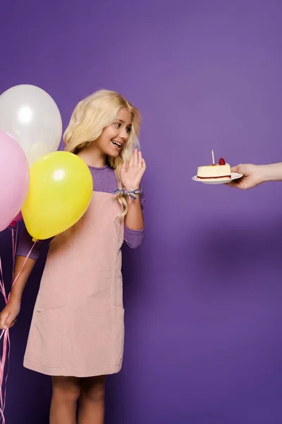 Vista recortada de mujer dando plato con pastel de cumpleaños a niño sonriente con globos sobre fondo púrpura - foto de stock