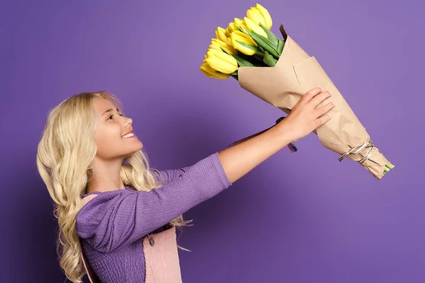 Side view of smiling kid holding bouquet of tulips on purple background — Stock Photo