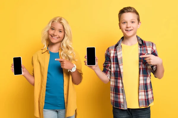 Niños sonrientes señalando con los dedos a los teléfonos inteligentes sobre fondo amarillo - foto de stock