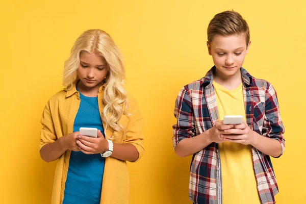 Niños sonrientes y lindos usando teléfonos inteligentes sobre fondo amarillo - foto de stock