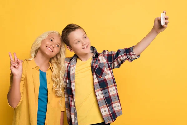 Smiling kid showing peace gesture and friend taking selfie on yellow background — Stock Photo