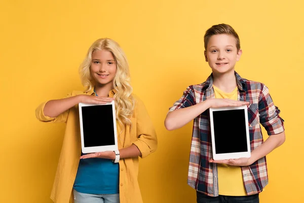 Niños sonrientes sosteniendo tabletas digitales con espacio de copia sobre fondo amarillo - foto de stock