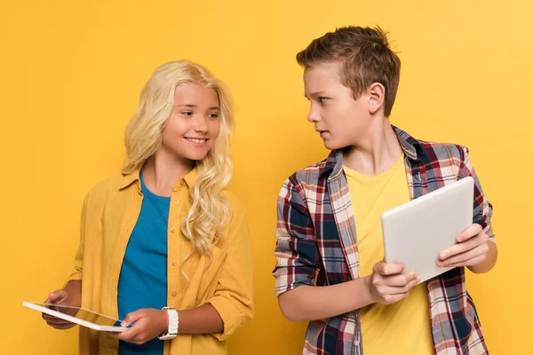 Улыбающийся ребенок смотрит на цифровой планшет своей подруги на желтом фоне — стоковое фото
