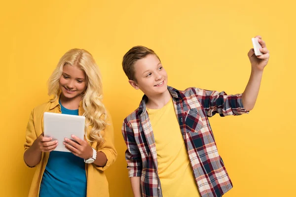 Niño sonriente tomando selfie y su amigo usando tableta digital sobre fondo amarillo - foto de stock