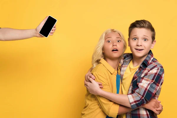 Cropped view of woman holding smartphone and scared kids hugging on yellow background — Stock Photo