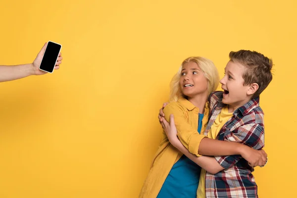Vista recortada de la mujer sosteniendo el teléfono inteligente y los niños asustados abrazando y gritando sobre el fondo amarillo - foto de stock