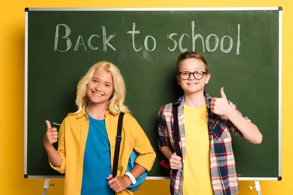 Écoliers souriants montrant pouces vers le haut près de tableau noir avec retour à l'école lettrage — Photo de stock