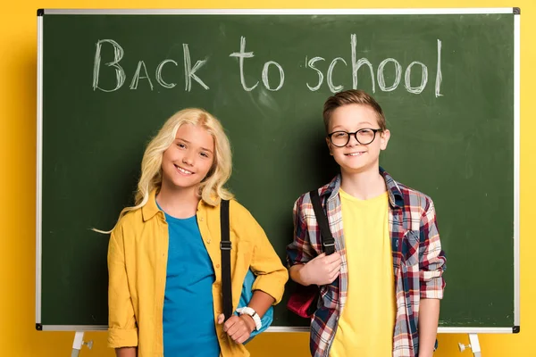 Smiling schoolkids with backpacks standing near chalkboard with back to school lettering — Stock Photo