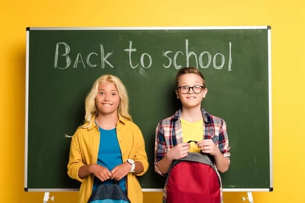 Smiling schoolkids with backpacks standing near chalkboard with back to school lettering — Stock Photo