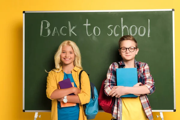 Smiling schoolkids holding books and standing near chalkboard with back to school lettering — Stock Photo