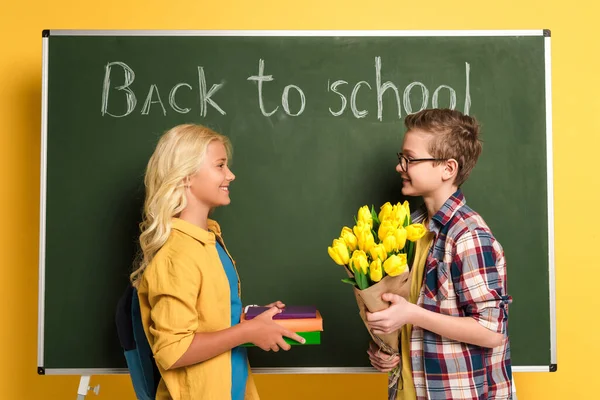 Vue latérale d'écoliers souriants tenant des livres et un bouquet près du tableau noir avec lettrage de retour à l'école — Photo de stock