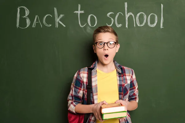 Sorprendido colegial con libros de pie cerca de pizarra con letras de vuelta a la escuela - foto de stock