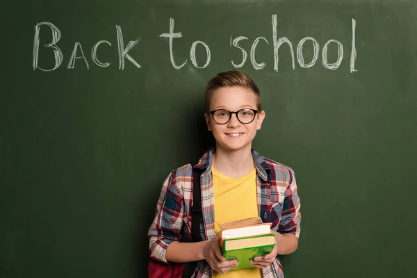 Écolier souriant avec des livres debout près du tableau noir avec lettrage de retour à l'école — Photo de stock