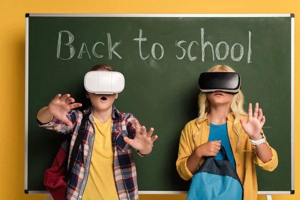 Shocked schoolkids with Virtual reality headsets standing near chalkboard with back to school lettering — Stock Photo