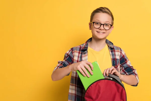 Écolier souriant mettre le livre dans un sac à dos sur fond jaune — Photo de stock