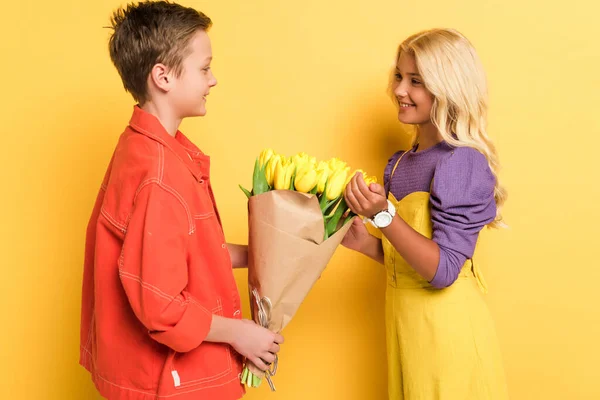 Side view of smiling boy giving bouquet to cute friend on yellow background — Stock Photo