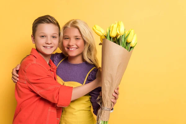 Улыбающийся мальчик обнимает друга с букетом на желтом фоне — стоковое фото