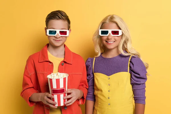 Улыбающиеся дети в 3D очках, держащие попкорн на желтом фоне — стоковое фото