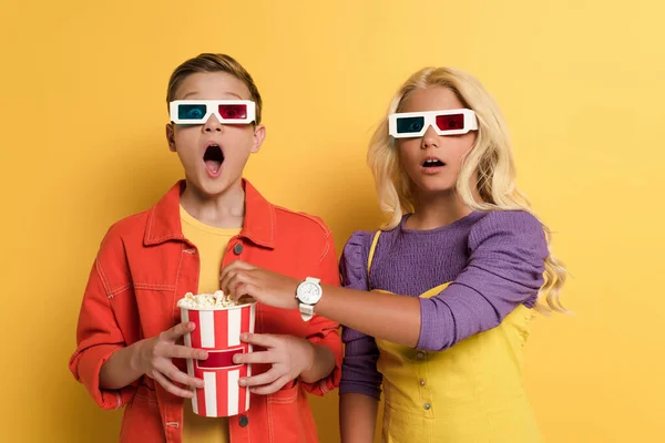 Enfants choqués avec des lunettes 3D tenant pop-corn et regardant la caméra sur fond jaune — Photo de stock