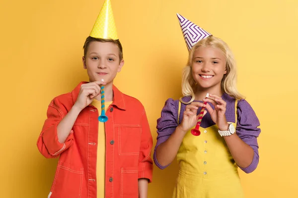Niños sonrientes con gorras de fiesta sosteniendo cuernos de fiesta sobre fondo amarillo - foto de stock