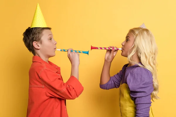 Side view of kids with party caps blowing in party horns on yellow background — Stock Photo