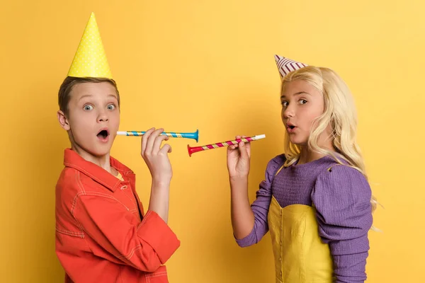 Niños impactados con gorras de fiesta con cuernos de fiesta sobre fondo amarillo - foto de stock