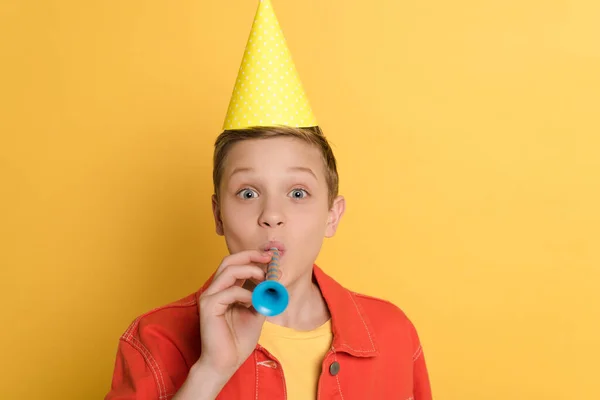 Kind in Partymütze bläst in Partyhorn auf gelbem Hintergrund — Stockfoto