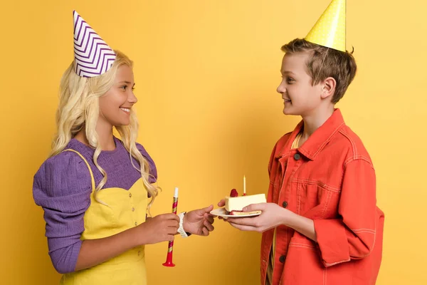 Vista lateral del niño dando plato con pastel de cumpleaños a un amigo sonriente con gorra de fiesta sobre fondo amarillo - foto de stock