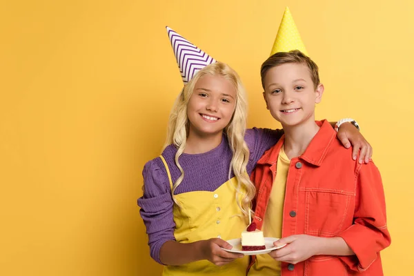 Enfants souriants avec casquettes de fête tenant plaque avec gâteau d'anniversaire et câlins sur fond jaune — Photo de stock